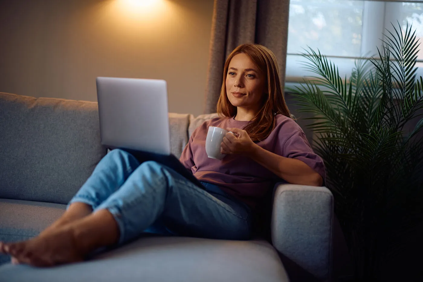 A woman relaxes on her sofa with a laptop and coffee during an online session